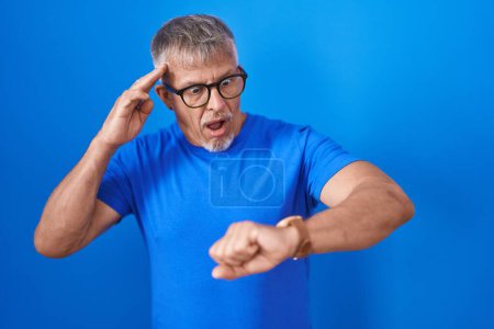 Téléchargez les photos : Homme hispanique avec les cheveux gris debout sur fond bleu regardant le temps de la montre inquiet, peur de se faire tard - en image libre de droit
