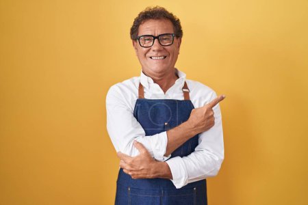 Téléchargez les photos : Homme hispanique du Moyen Âge portant tablier de cuisine professionnel avec un grand sourire sur le visage, pointant avec la main et le doigt vers le côté regardant la caméra. - en image libre de droit