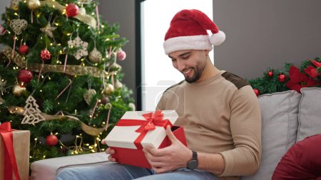 Foto de Joven árabe hombre desembalaje regalo sentado en sofá por árbol de Navidad en casa - Imagen libre de derechos