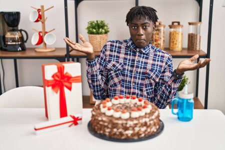 Photo for African man with dreadlocks celebrating birthday holding big chocolate cake clueless and confused expression with arms and hands raised. doubt concept. - Royalty Free Image