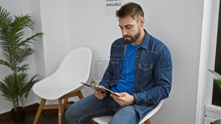 Young hispanic man sitting on chair reading document on clipboard at waiting room