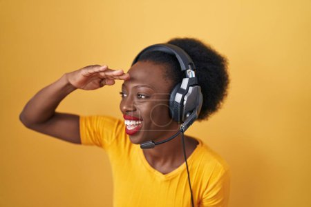 Photo for African woman with curly hair standing over yellow background wearing headphones very happy and smiling looking far away with hand over head. searching concept. - Royalty Free Image