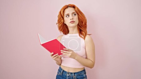 Photo for Young redhead woman reading book with serious expression over isolated pink background - Royalty Free Image