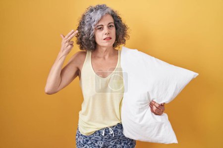 Foto de Mujer de mediana edad con el pelo gris usando pijama abrazando almohada disparando y matándose señalando la mano y los dedos a la cabeza como arma, gesto suicida. - Imagen libre de derechos