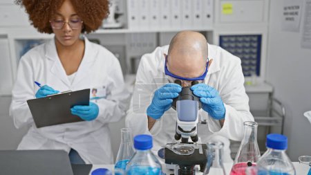 Photo for Inside the buzzing lab, two committed scientists, man and woman, analysis partners, seated together, gloved hands busy writing on clipboard, eyes glued to the microscope in serious research - Royalty Free Image