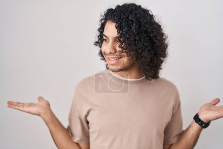 Téléchargez les photos : Homme hispanique aux cheveux bouclés debout sur fond blanc souriant montrant les paumes ouvertes des deux mains, présentant et comparant la publicité et l'équilibre - en image libre de droit