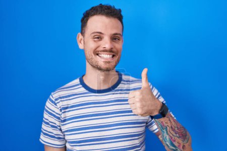 Téléchargez les photos : Jeune homme hispanique debout sur fond bleu faisant des pouces heureux geste avec la main. approuvant l'expression regardant la caméra montrant le succès. - en image libre de droit