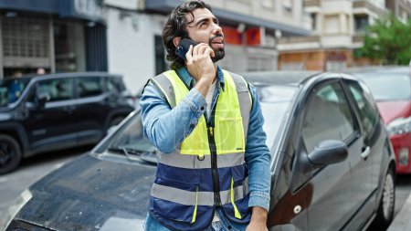 Photo for Young hispanic man standing by car wearing reflective vest talking on smartphone at street - Royalty Free Image