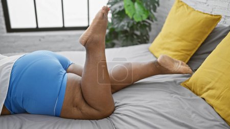 Téléchargez les photos : Belle femme afro-américaine confortablement relaxante au lit, jambes mobiles dans la chambre confortable matin - en image libre de droit