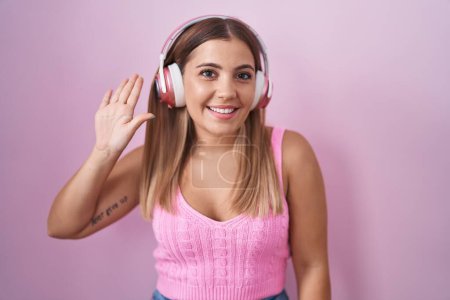 Téléchargez les photos : Jeune femme blonde écoutant de la musique à l'aide d'écouteurs renonçant à dire bonjour heureux et souriant, geste de bienvenue amical - en image libre de droit