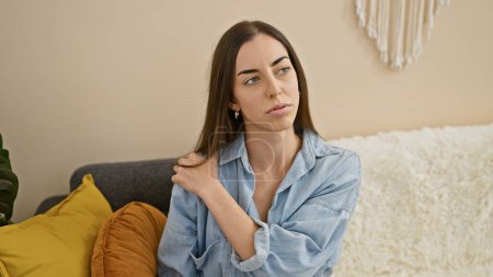 Photo for Beautiful young hispanic woman, sitting in cozy indoor living room, suffering intense back pain, stress etched on her face, doubting what to do next. - Royalty Free Image