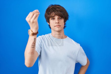 Foto de Joven hispano de pie sobre fondo azul haciendo gesto italiano con la mano y los dedos expresión confiada - Imagen libre de derechos