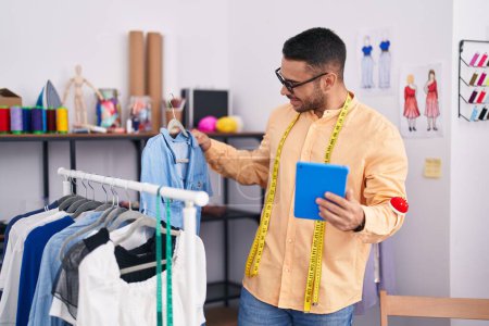 Téléchargez les photos : Jeune homme hispanique tailleur tenant des vêtements sur support à l'aide de pavé tactile au magasin de tailleur - en image libre de droit