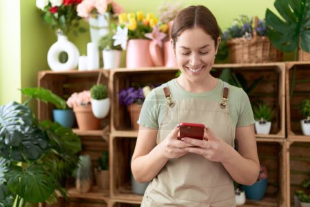 Téléchargez les photos : Jeune belle femme fleuriste souriant confiant en utilisant un smartphone à la boutique de fleurs - en image libre de droit