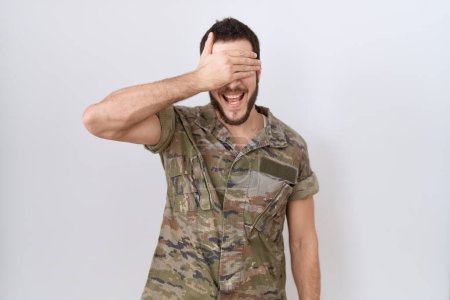 Téléchargez les photos : Jeune homme hispanique portant uniforme de camouflage de l'armée souriant et riant avec la main sur le visage couvrant les yeux pour la surprise. concept aveugle. - en image libre de droit
