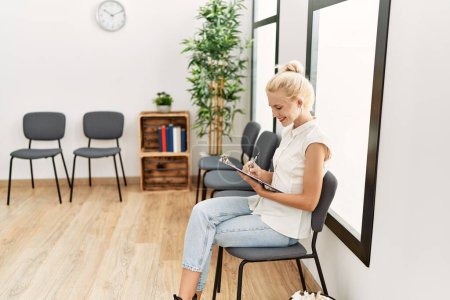 Téléchargez les photos : Jeune femme blonde utilisant un smartphone assis sur une chaise dans la salle d'attente - en image libre de droit