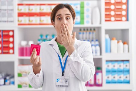 Foto de Mujer morena trabajando en farmacia sosteniendo condón cubriendo la boca con la mano, conmocionada y temerosa de equivocarse. expresión sorprendida - Imagen libre de derechos