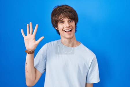 Photo for Hispanic young man standing over blue background showing and pointing up with fingers number five while smiling confident and happy. - Royalty Free Image