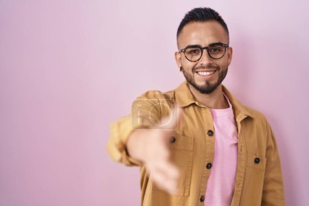 Foto de Joven hombre hispano de pie sobre fondo rosa sonriente amistoso ofreciendo apretón de manos como saludo y bienvenida. negocios exitosos. - Imagen libre de derechos