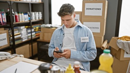 Photo for Handsome bearded young caucasian man volunteering at a local charity center, focused on his smartphone as he types out messages for donations, sitting indoors, surrounded by cardboard boxes - Royalty Free Image