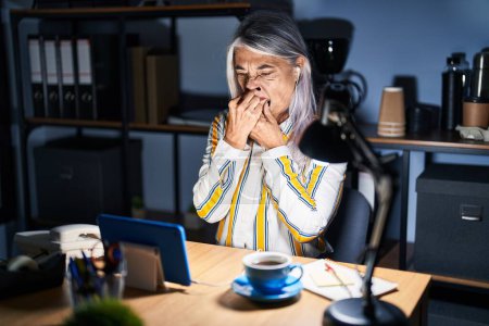 Foto de Mujer de mediana edad con el pelo gris trabajando en la oficina por la noche oliendo algo apestoso y asqueroso, olor intolerable, conteniendo la respiración con los dedos en la nariz. mal olor - Imagen libre de derechos