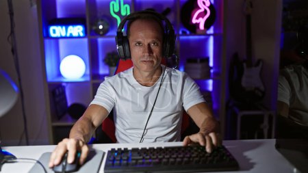 Téléchargez les photos : Bel homme d'âge moyen streaming jeu vidéo la nuit, assis sérieusement concentré dans sa salle de jeux cyber, entièrement équipé avec la technologie futuriste - en image libre de droit