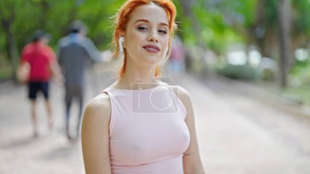 Foto de Mujer pelirroja joven con ropa deportiva y auriculares sonriendo en el parque - Imagen libre de derechos