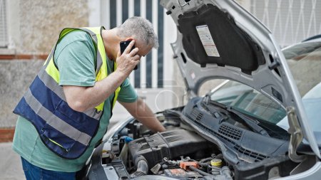 Foto de Hombre de pelo gris de mediana edad hablando en el teléfono inteligente con el seguro de reparación de coches en la calle - Imagen libre de derechos