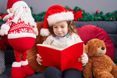 Foto de Adorable rubio niño leyendo libro sentado en el sofá por la decoración de Navidad en casa - Imagen libre de derechos