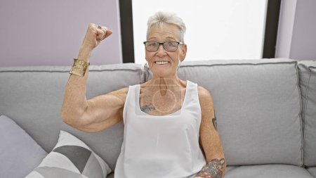 Téléchargez les photos : Femme âgée aux cheveux gris confiante fléchissant joyeusement ses biceps sur le canapé du salon à la maison, arborant un grand sourire joyeux et une attitude cool et positive - en image libre de droit