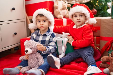 Photo for Two kids holding gift and teddy bear sitting on floor by christmas tree at home - Royalty Free Image