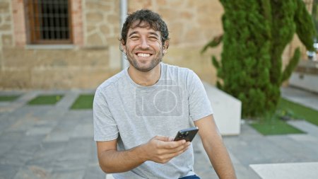 Téléchargez les photos : Jeune homme beau et confiant, envoyant joyeusement des SMS sur son smartphone, assis avec une expression décontractée dans le parc de la ville, profitant d'une journée ensoleillée en plein air. - en image libre de droit