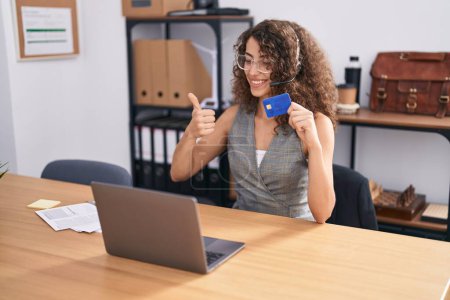 Photo for Hispanic woman with curly hair wearing call center agent headset holding credit card smiling happy and positive, thumb up doing excellent and approval sign - Royalty Free Image