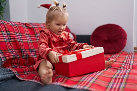 Foto de Adorable chica rubia desempacar regalo de Navidad sentado en el sofá en casa - Imagen libre de derechos