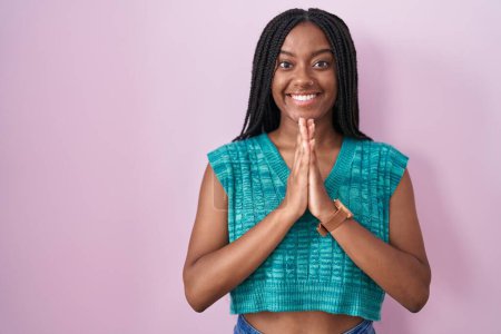 Foto de Joven afroamericano con trenzas de pie sobre fondo rosa rezando con las manos juntas pidiendo perdón sonriendo confiado. - Imagen libre de derechos