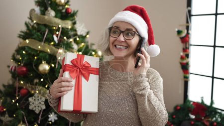 Foto de Mujer de mediana edad con el pelo gris hablando en el teléfono inteligente de pie junto al árbol de Navidad en casa - Imagen libre de derechos