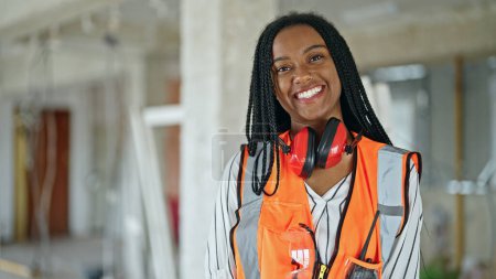 Téléchargez les photos : Femme afro-américaine constructeur souriant confiance en portant des écouteurs sur le chantier de construction - en image libre de droit