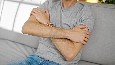 Téléchargez les photos : Jeune homme hispanique se gratter le bras pour démangeaisons assis sur le canapé à la maison - en image libre de droit