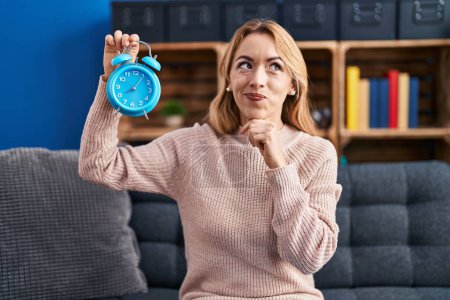 Photo for Hispanic woman holding alarm clock serious face thinking about question with hand on chin, thoughtful about confusing idea - Royalty Free Image