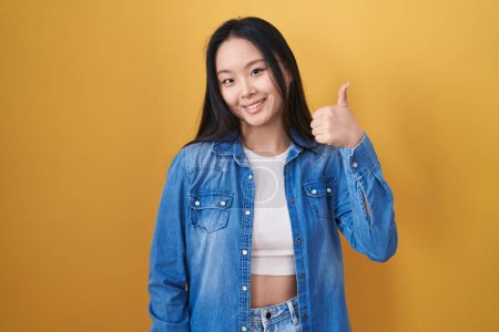 Téléchargez les photos : Jeune femme asiatique debout sur fond jaune faisant des pouces heureux geste avec la main. approuvant l'expression regardant la caméra montrant le succès. - en image libre de droit