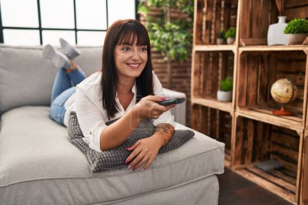 Téléchargez les photos : Jeune belle femme hispanique regarder la télévision couché sur le canapé à la maison - en image libre de droit