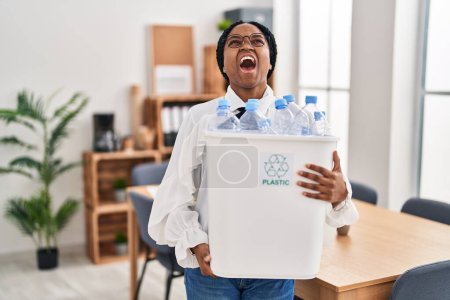 Foto de Mujer afroamericana trabajando en la oficina sosteniendo botella de plástico para reciclar enojado y loco gritando frustrado y furioso, gritando con ira mirando hacia arriba. - Imagen libre de derechos