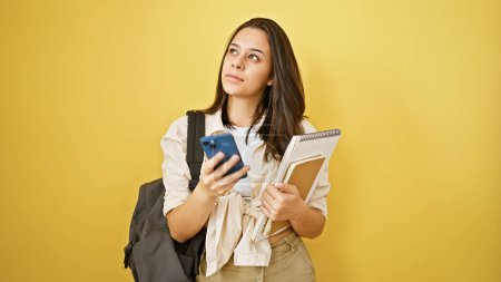Foto de Mujer hispana joven pensativa, una estudiante inteligente, sosteniendo libro y teléfono, dudando, aislada sobre fondo amarillo - Imagen libre de derechos