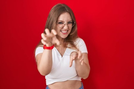 Téléchargez les photos : Jeune femme caucasienne debout sur fond rouge souriant drôle faisant griffe geste comme chat, expression agressive et sexy - en image libre de droit