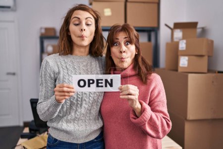 Photo for Two women working at small business ecommerce holding open banner making fish face with mouth and squinting eyes, crazy and comical. - Royalty Free Image