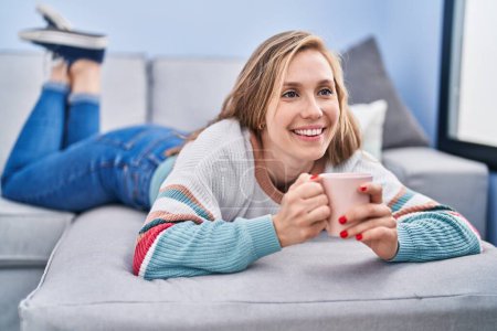 Foto de Mujer rubia joven bebiendo café tumbado en el sofá en casa - Imagen libre de derechos