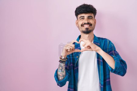 Téléchargez les photos : Jeune homme hispanique avec la barbe debout sur fond rose souriant dans l'amour faisant forme de symbole de coeur avec les mains. concept romantique. - en image libre de droit
