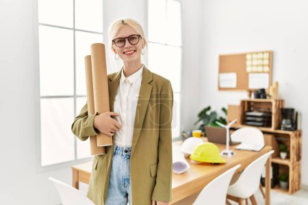 Photo for Young blonde woman architect smiling confident holding blueprints at office - Royalty Free Image
