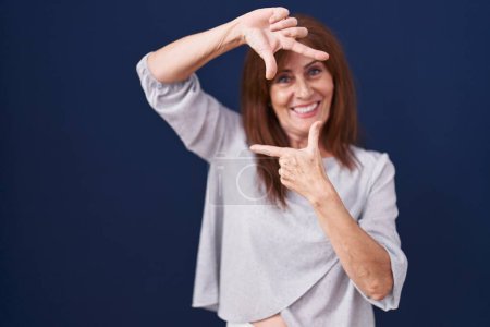 Téléchargez les photos : Femme d'âge moyen debout sur fond bleu souriant faisant cadre avec les mains et les doigts avec le visage heureux. concept de créativité et photographie. - en image libre de droit