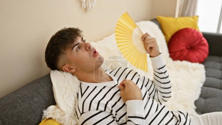 Foto de Guapo joven hispano sudando por el calor, encuentra consuelo en la brisa fresca de un ventilador de mano, sentado en un acogedor sofá en la sala de estar en casa, la preocupación grabada en su cara. - Imagen libre de derechos